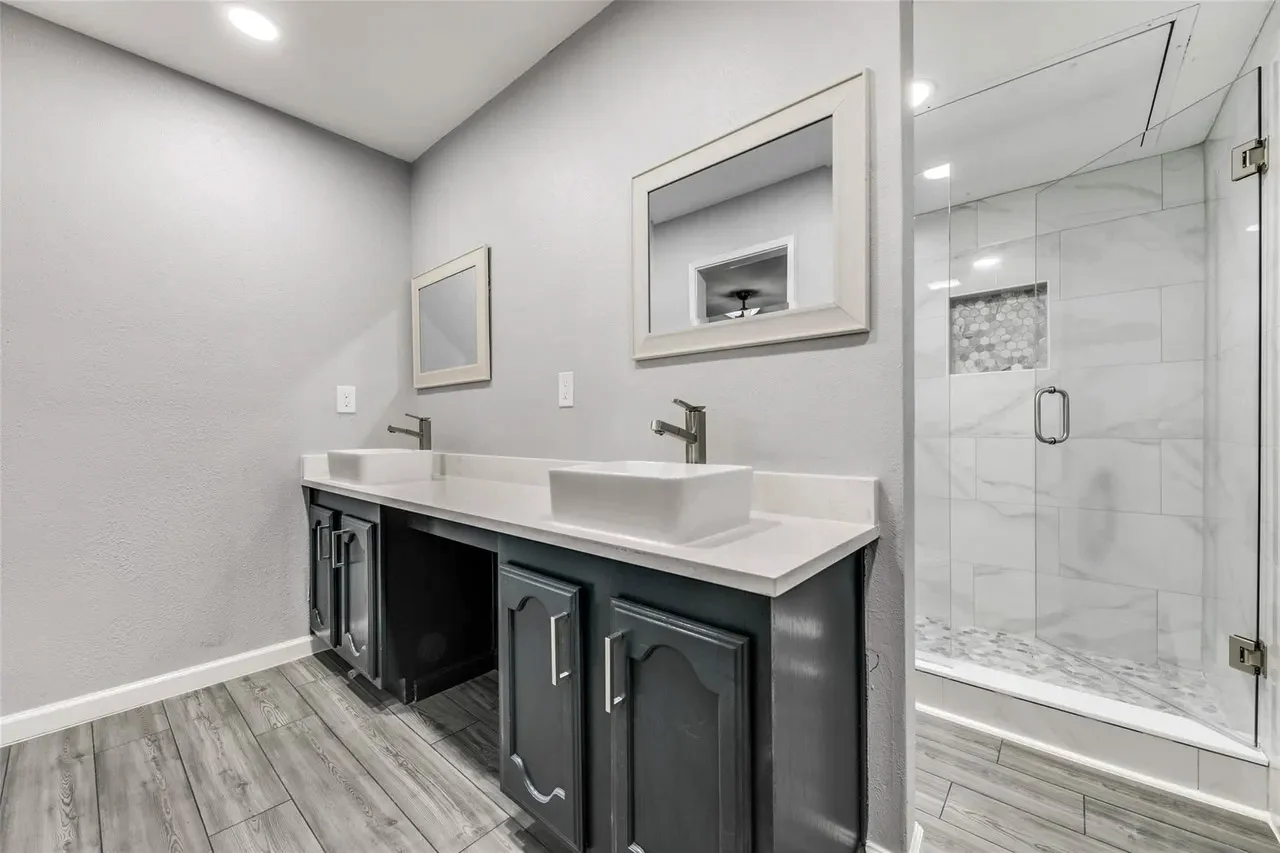 Primary bathroom with a dark double vanity, white counters, frameless glass shower, marble-look tile, and chrome fixtures in Dallas, Texas.