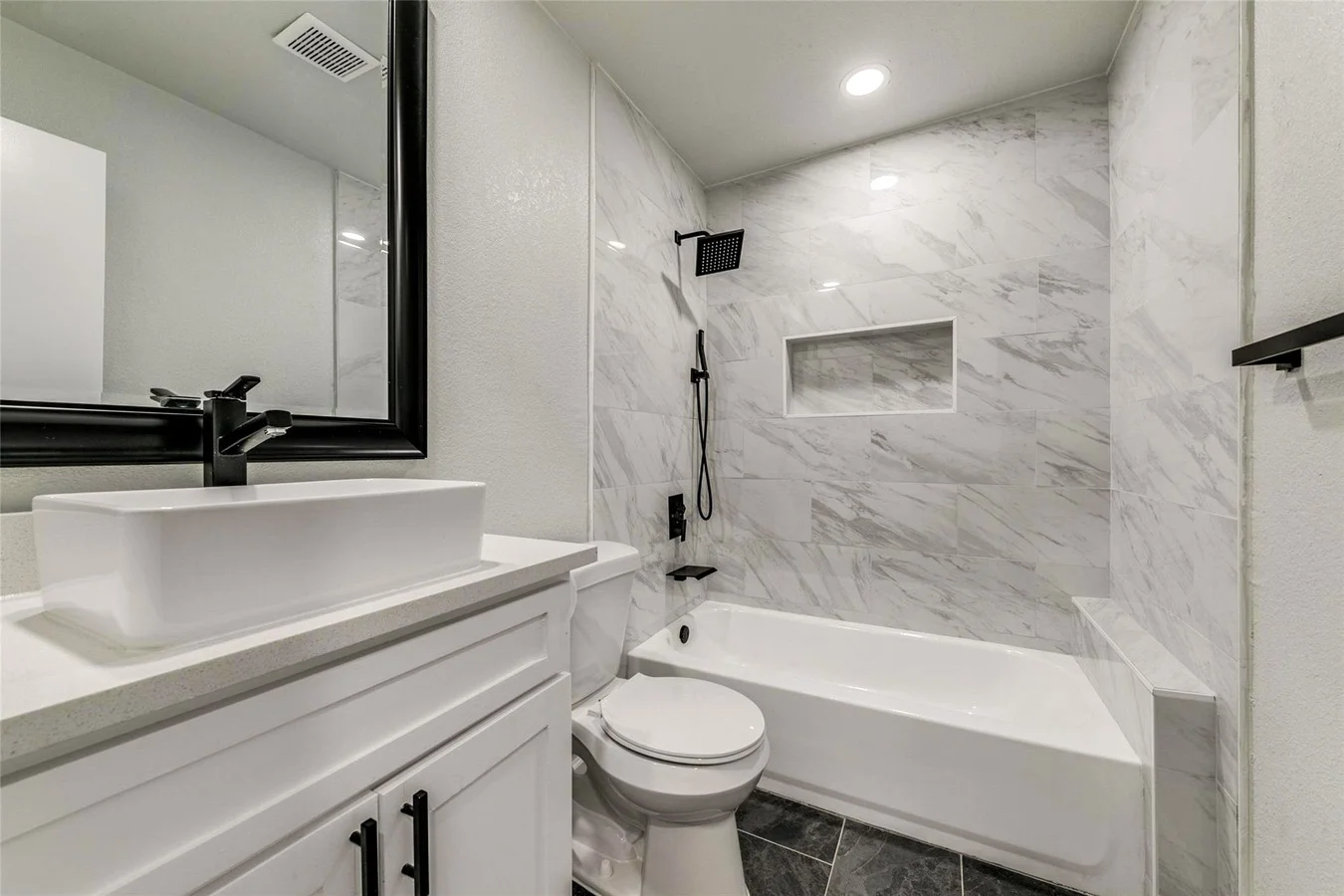 Guest bathroom with a marble-look tub surround, white vessel sink, black fixtures, gray vanity, and recessed lighting in Plano, Texas.