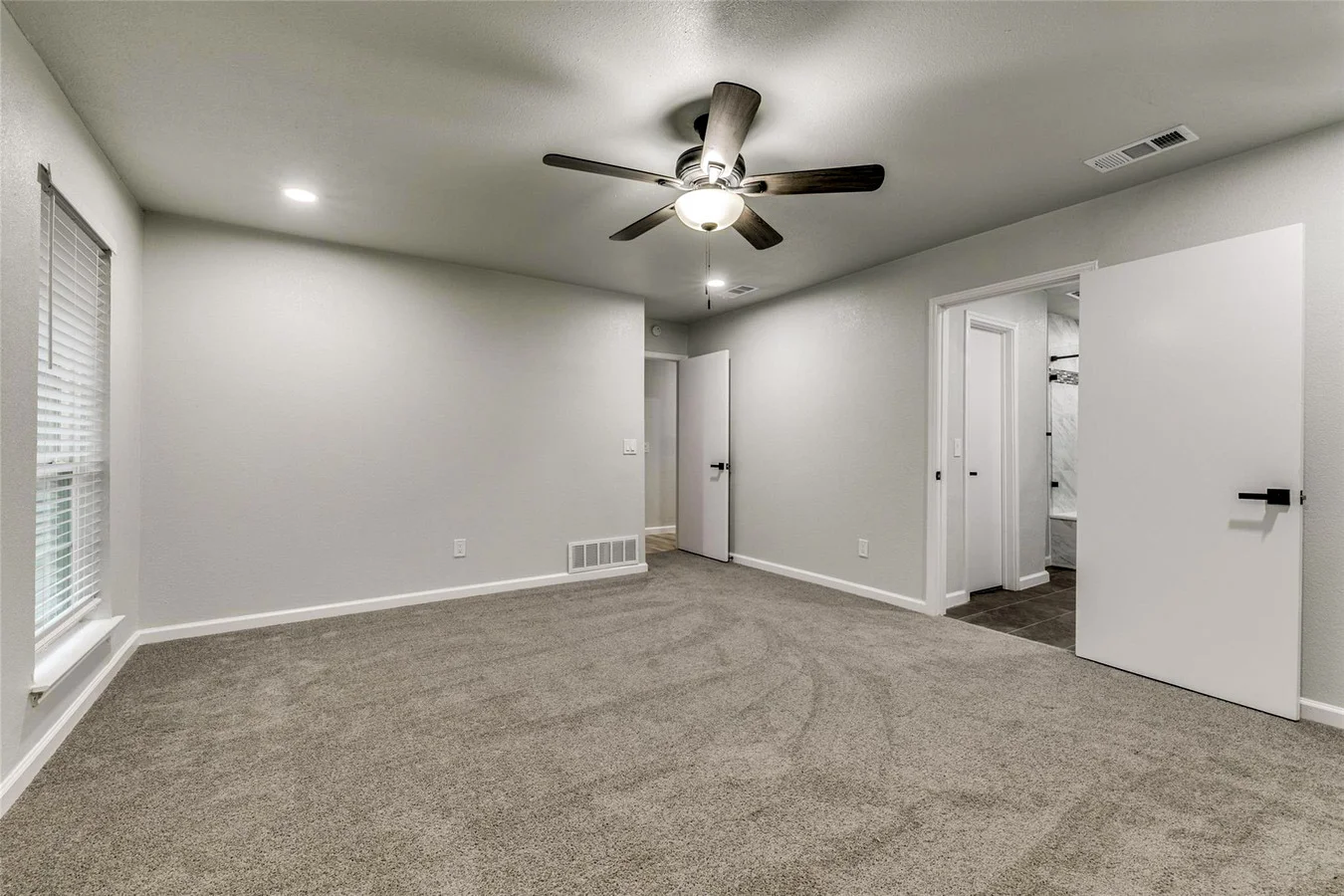 Primary bedroom with new gray carpet, light gray walls, ceiling fan, white trim, and adjacent bathroom access in Plano, Texas.